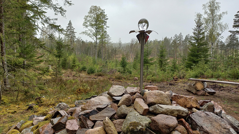 Foto på en stolpe med en kristallkula på. Konstverket är placerat i ett stenröse i skogen.